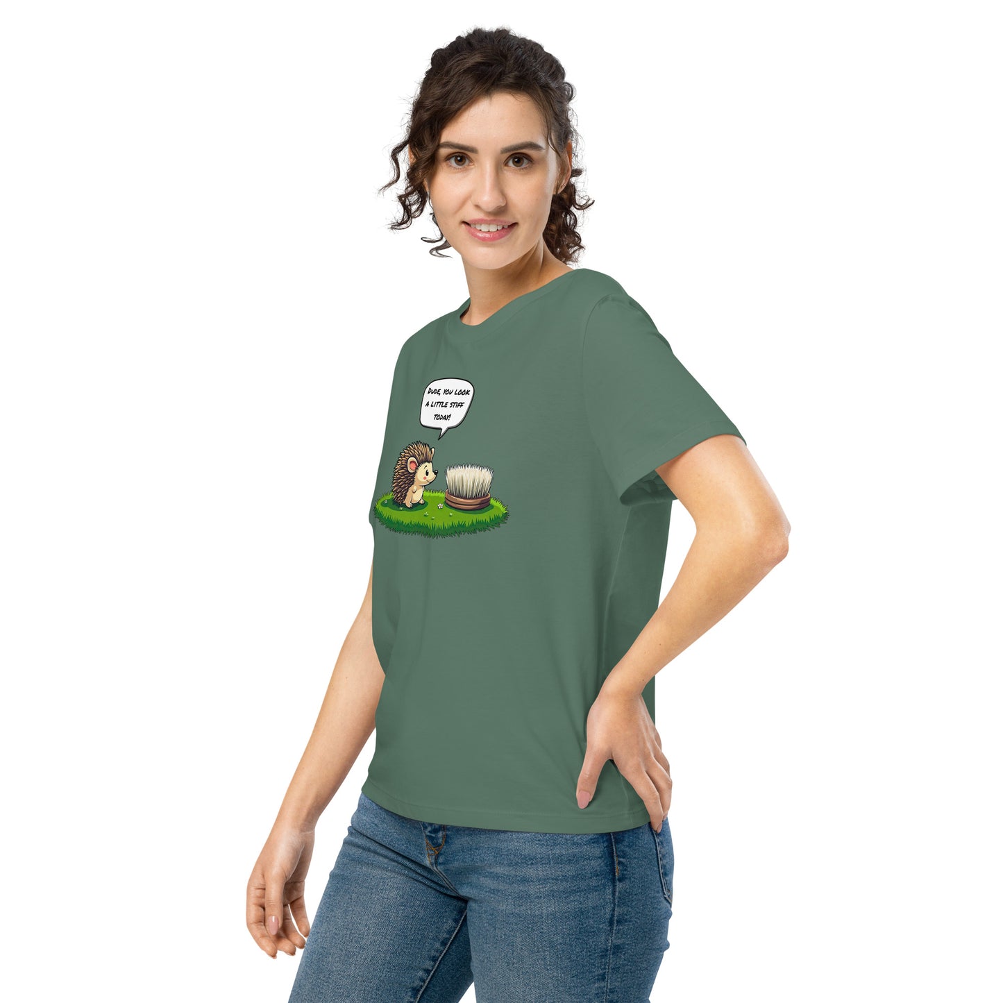 Product mockup. A happy woman wearing a green organic cotton t-shirt featuring a playful cartoon illustration of a hedgehog standing on green grass beside a scrubbing brush. The hedgehog has a speech bubble that says, 'DUDE, YOU LOOK A LITTLE STIFF TODAY!'