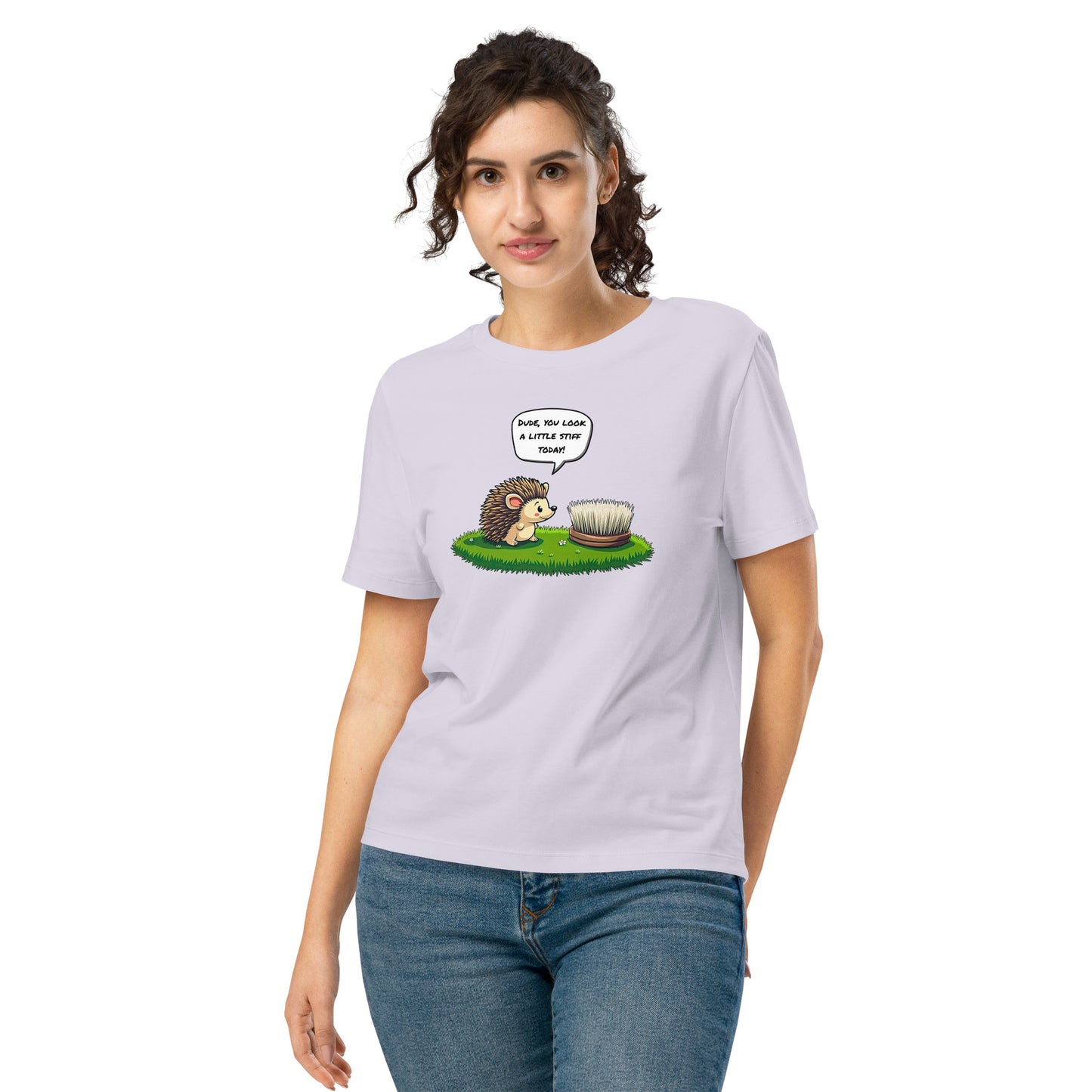 Product mockup. A happy woman wearing a lavender organic cotton t-shirt featuring a playful cartoon illustration of a hedgehog standing on green grass beside a scrubbing brush. The hedgehog has a speech bubble that says, 'DUDE, YOU LOOK A LITTLE STIFF TODAY!'