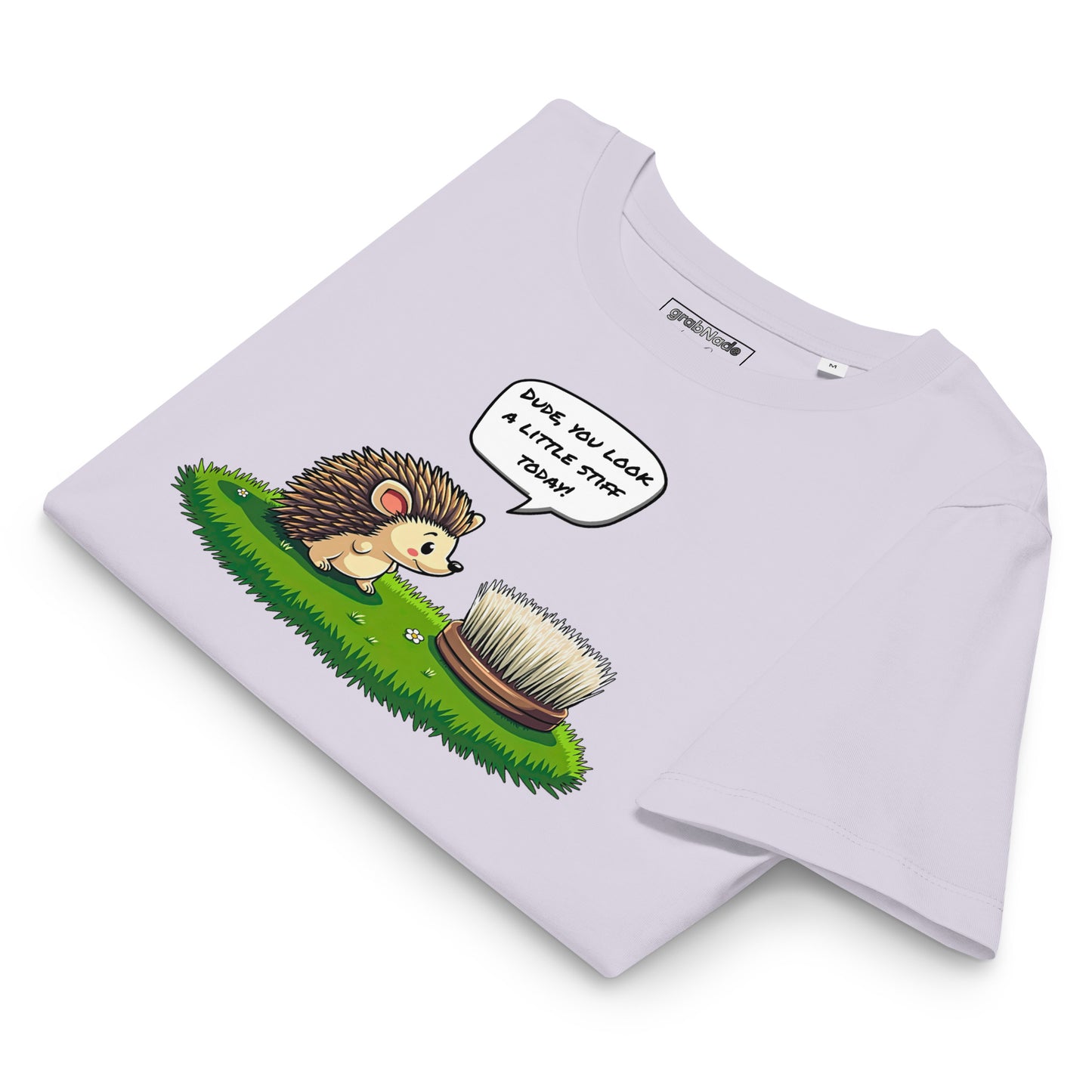 Product mockup. A folded lavender t-shirt made from organic cotton, featuring a playful illustration on the front. The design shows a cute cartoon hedgehog standing on green grass, looking at a stiff scrubbing brush, with a speech bubble that reads, 'DUDE, YOU LOOK A LITTLE STIFF TODAY!' The 'grabNade' brand label is visible inside the neckline.
