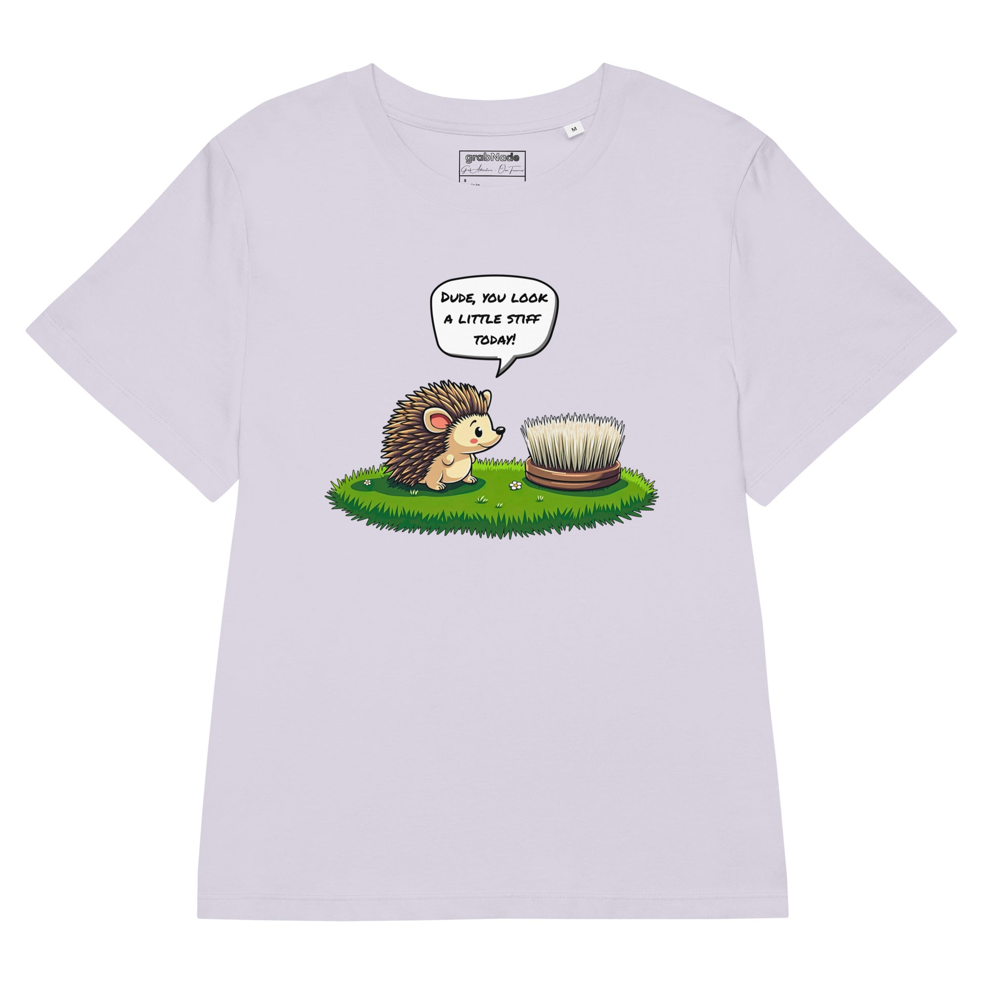 Product mockup. A flat layed out lavender t-shirt made from organic cotton, featuring a playful illustration on the front. The design shows a cute cartoon hedgehog standing on green grass, looking at a stiff scrubbing brush, with a speech bubble that reads, 'DUDE, YOU LOOK A LITTLE STIFF TODAY!' The 'grabNade' brand label is visible inside the neckline.