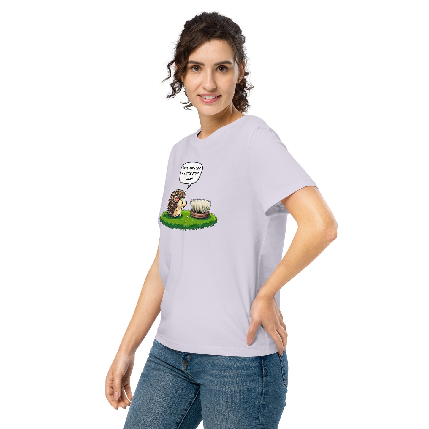 Product mockup. A happy woman wearing a lavender organic cotton t-shirt featuring a playful cartoon illustration of a hedgehog standing on green grass beside a scrubbing brush. The hedgehog has a speech bubble that says, 'DUDE, YOU LOOK A LITTLE STIFF TODAY!'