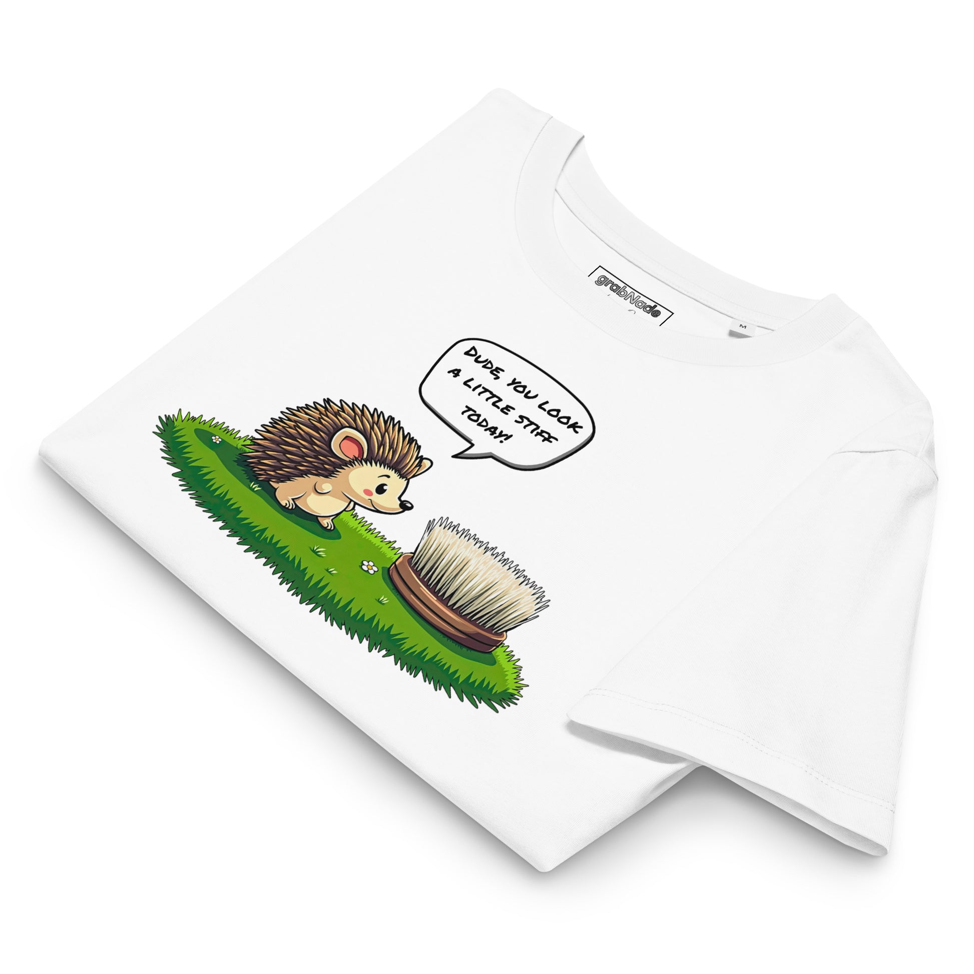Product mockup. A folded white t-shirt made from organic cotton, featuring a playful illustration on the front. The design shows a cute cartoon hedgehog standing on green grass, looking at a stiff scrubbing brush, with a speech bubble that reads, 'DUDE, YOU LOOK A LITTLE STIFF TODAY!' The 'grabNade' brand label is visible inside the neckline.