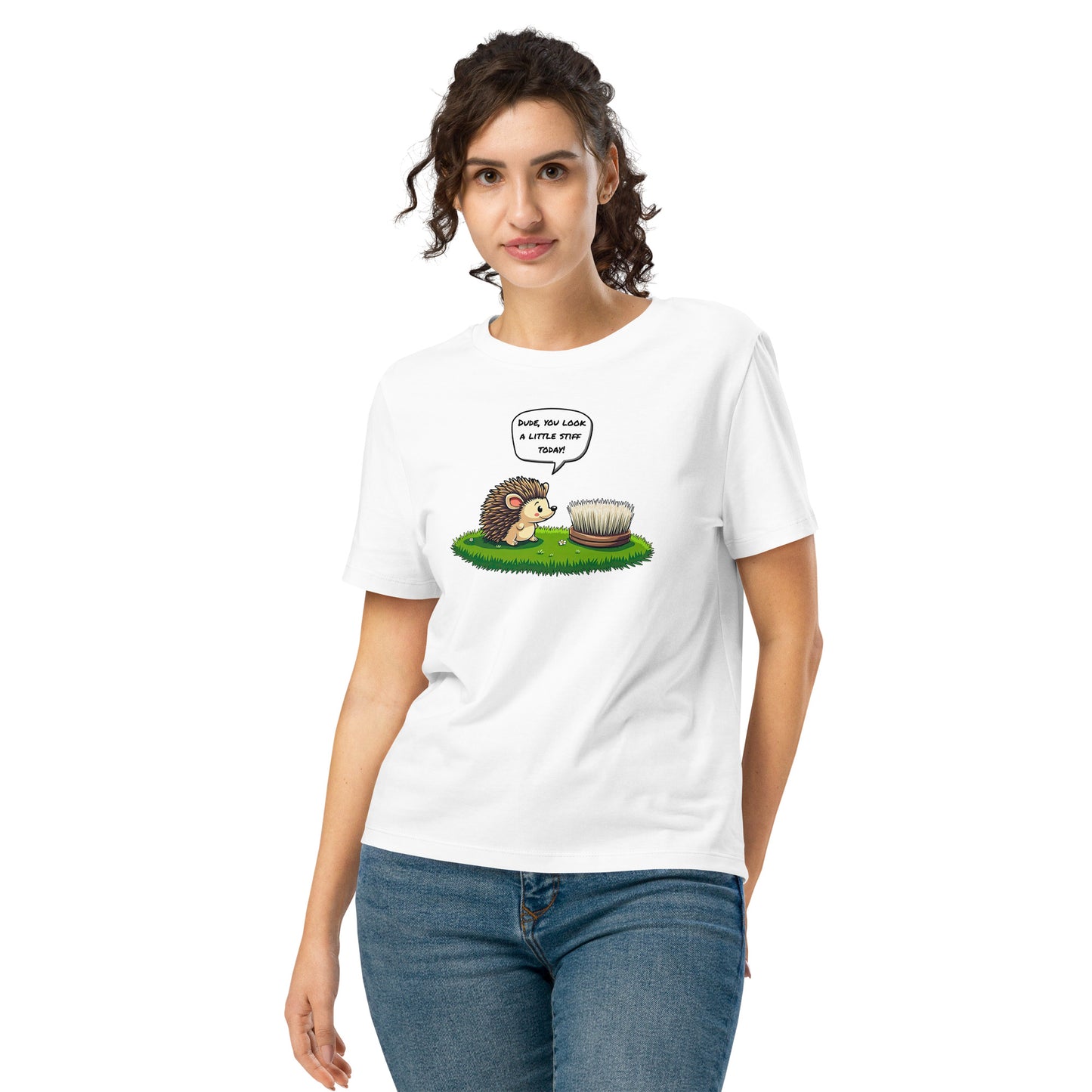 Product mockup. A happy woman wearing a white organic cotton t-shirt featuring a playful cartoon illustration of a hedgehog standing on green grass beside a scrubbing brush. The hedgehog has a speech bubble that says, 'DUDE, YOU LOOK A LITTLE STIFF TODAY!'