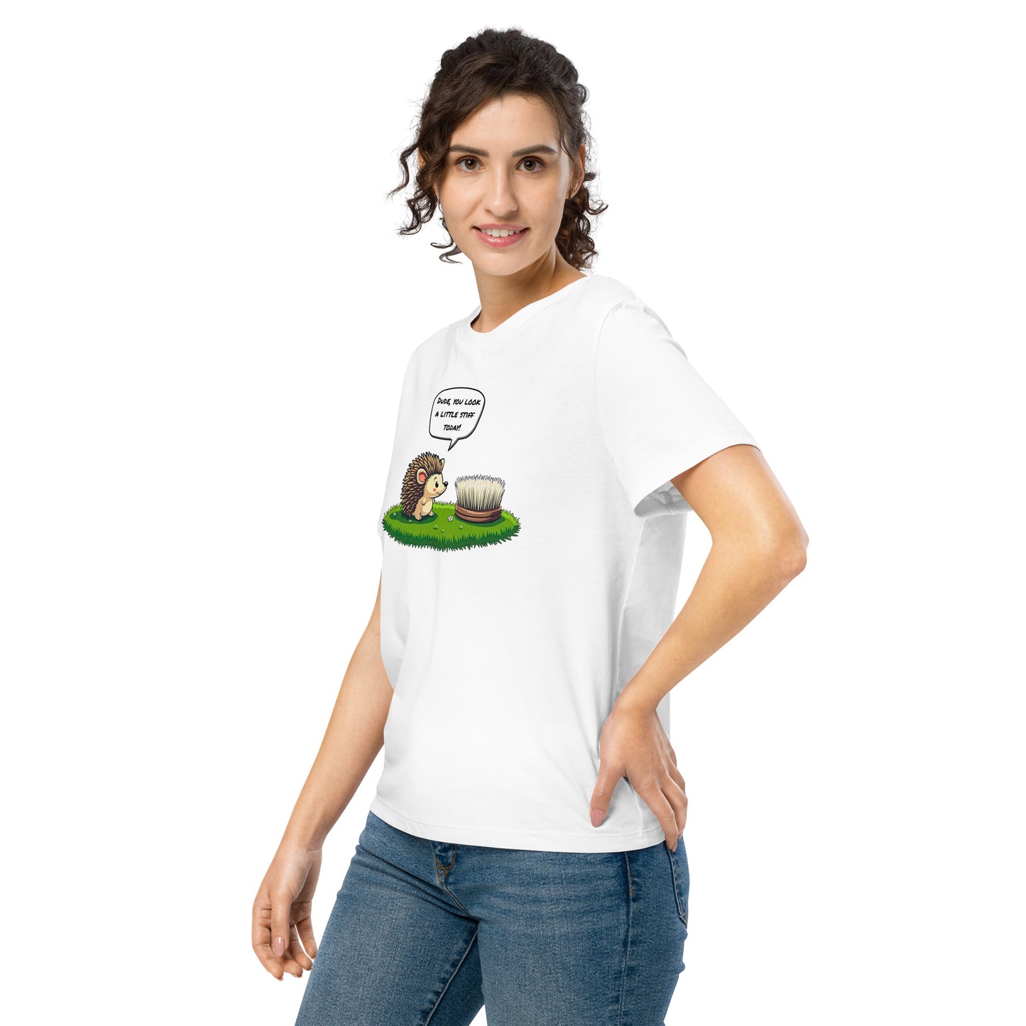 Product mockup. A happy woman wearing a white organic cotton t-shirt featuring a playful cartoon illustration of a hedgehog standing on green grass beside a scrubbing brush. The hedgehog has a speech bubble that says, 'DUDE, YOU LOOK A LITTLE STIFF TODAY!'