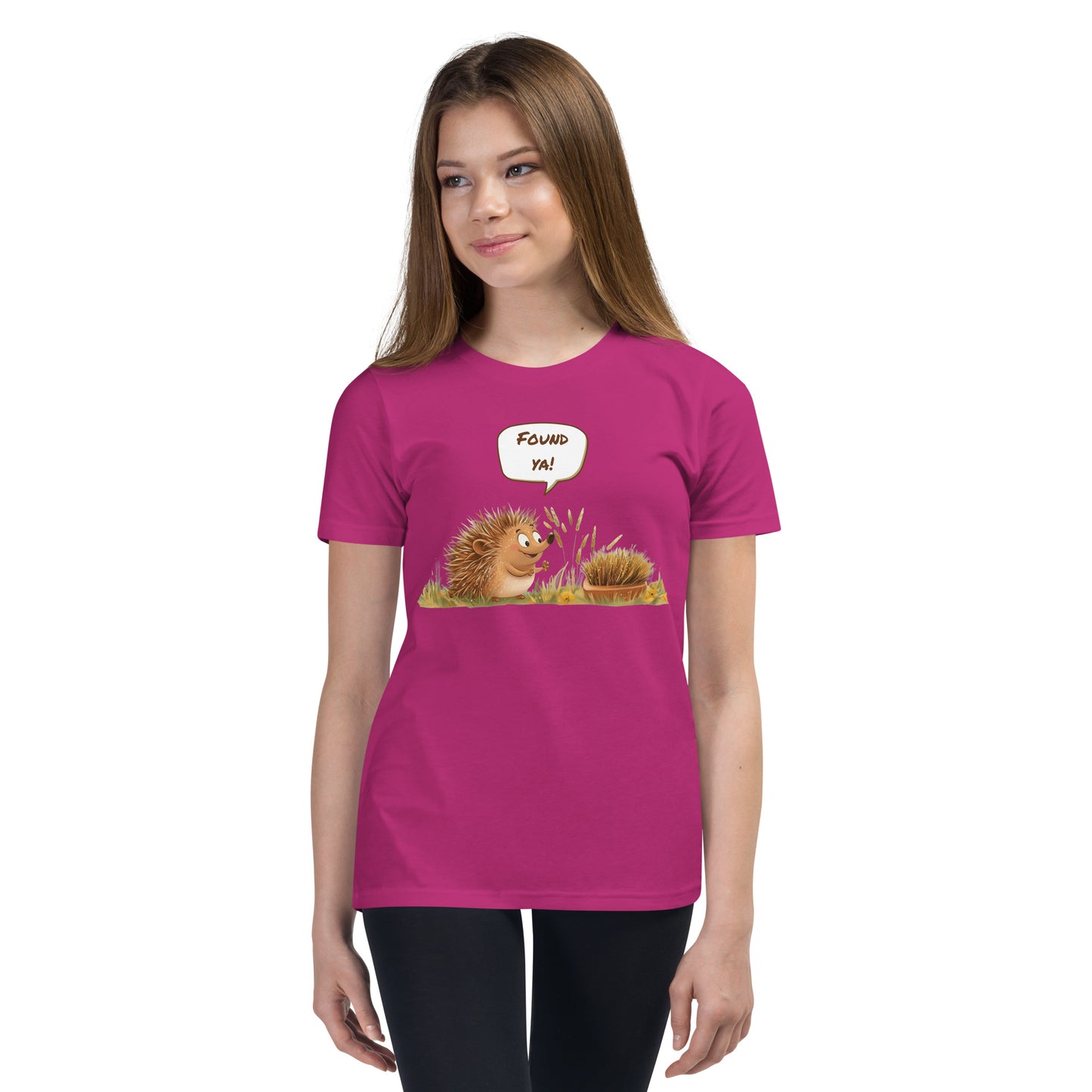Product mockup. Ultra-soft premium youth pink t-shirt featuring cute cartoon hedgehog with speech bubble saying 'FOUND YA!' over grassy ground, in front of a brush, modeled on young girl.