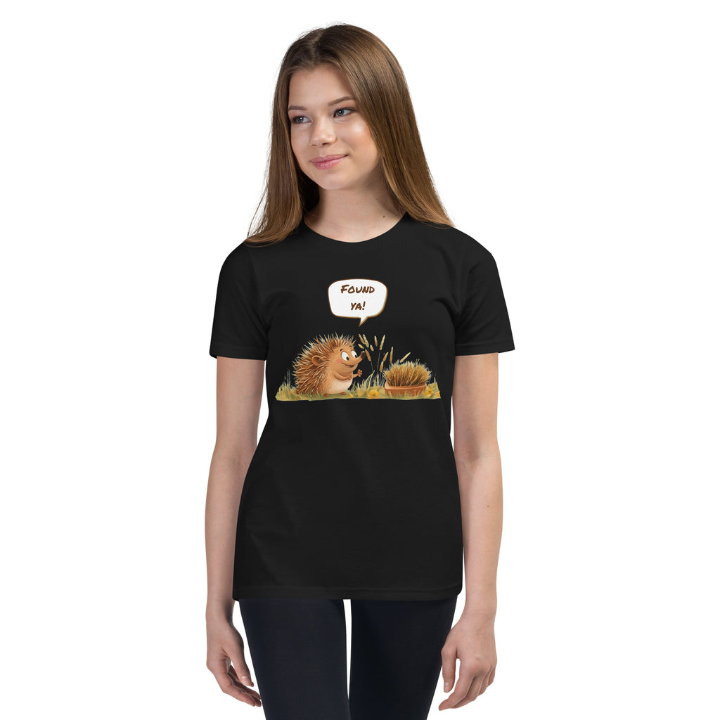 Product mockup. Ultra-soft premium youth black t-shirt featuring cute cartoon hedgehog with speech bubble saying 'FOUND YA!' over grassy ground, in front of a brush, modeled on young girl.