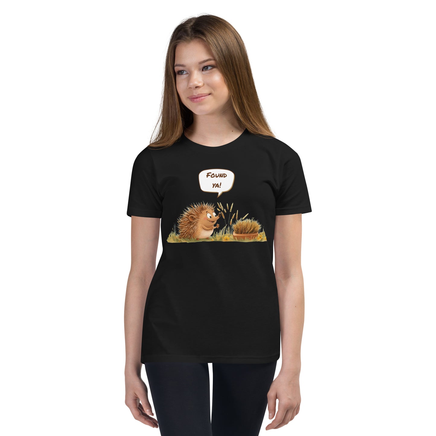 Product mockup. Ultra-soft premium youth black t-shirt featuring cute cartoon hedgehog with speech bubble saying 'FOUND YA!' over grassy ground, in front of a brush, modeled on young girl.