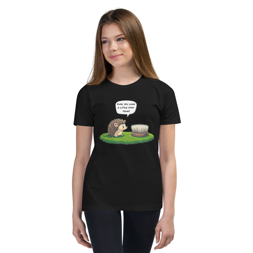 Product mockup. A young woman with long brown hair is facing the camera, wearing a black T-shirt and black pants. The T-shirt displays a cartoon hedgehog looking at a stiff brush on grass, with a speech bubble saying: Dude, you look a little stiff today!
