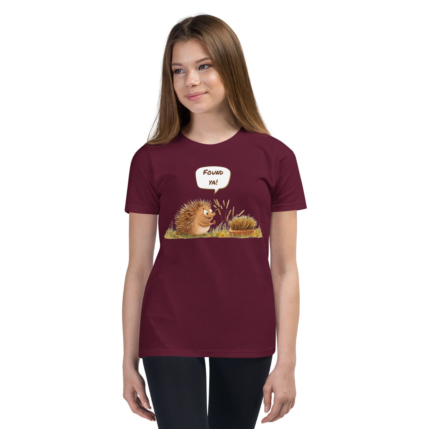 Product mockup. Ultra-soft premium youth maroon t-shirt featuring cute cartoon hedgehog with speech bubble saying 'FOUND YA!' over grassy ground, in front of a brush, modeled on young girl.