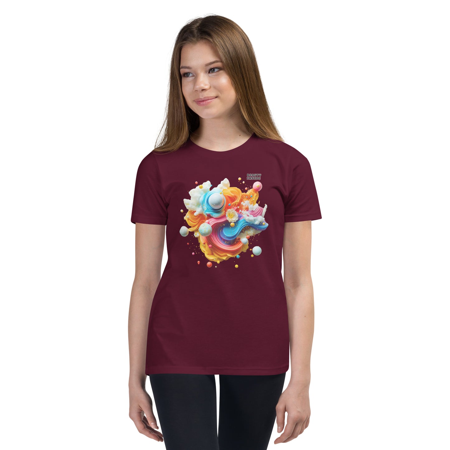 Product mockup. A young girl wearing a brown youth ultra-soft premium t-shirt with a colorful, abstract modern art design on the front. The design includes vibrant swirls and spheres in blue, pink, orange, yellow, and white, with a small 'BEAUTY INSIDE' text near the upper right part of the graphic. She is paired with black leggings and looking slightly to the side with a subtle smile.