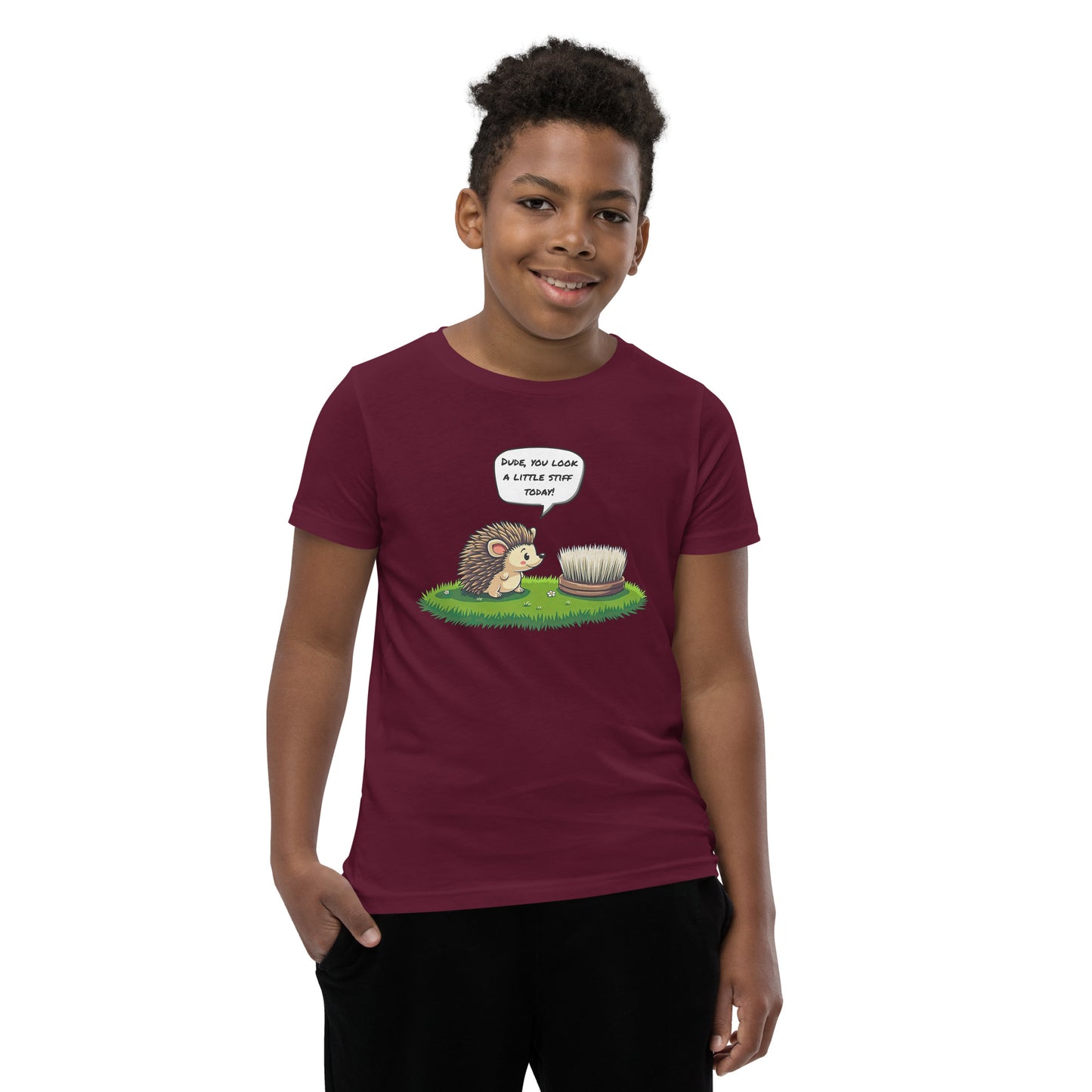 Product mockup. A boy wearing a maroon t-shirt featuring a playful cartoon illustration on the chest. The design shows a cute hedgehog standing on grass and looking at a scrubbing brush, with a speech bubble above the hedgehog that says, 'DUDE, YOU LOOK A LITTLE STIFF TODAY!