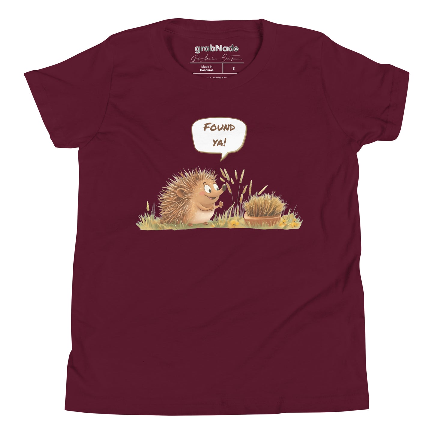 Product mockup. A smiling child wearing a light maroon t-shirt printed with a playful illustration of a cheerful hedgehog popping up from the grass and saying “Found ya!” in a speech bubble, beside a second hedgehog curled into a ball, all set against a clean studio background.