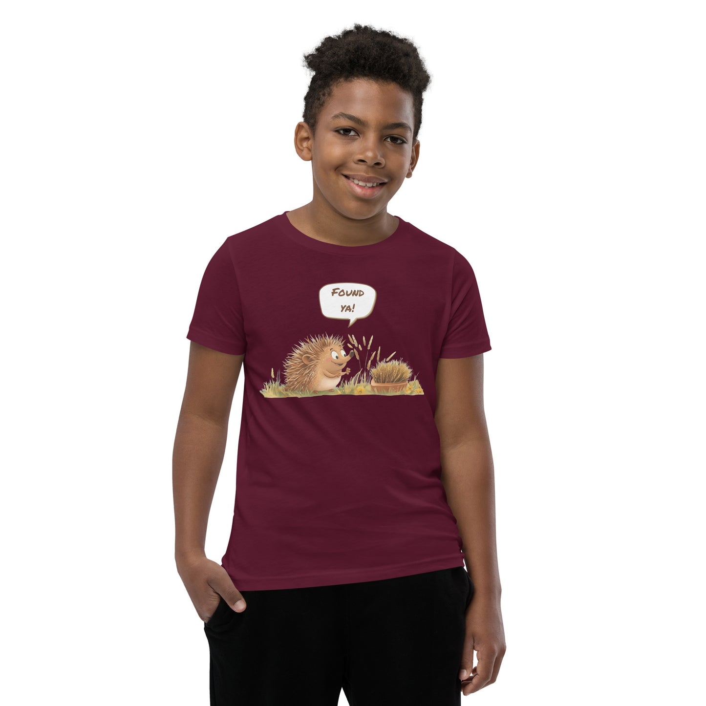 Product mockup. A smiling child wearing a maroon t-shirt printed with a playful illustration of a cheerful hedgehog popping up from the grass and saying “Found ya!” in a speech bubble, beside a second hedgehog curled into a ball, all set against a clean studio background.