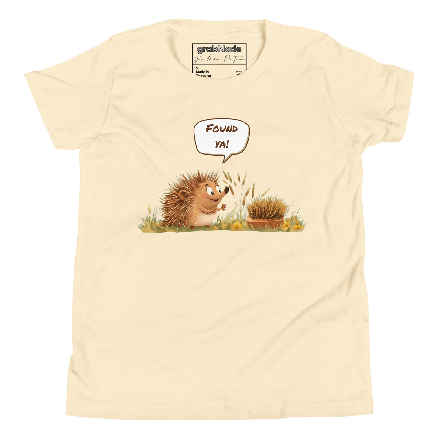 Product mockup. A smiling child wearing a light beige t-shirt printed with a playful illustration of a cheerful hedgehog popping up from the grass and saying “Found ya!” in a speech bubble, beside a second hedgehog curled into a ball, all set against a clean studio background.