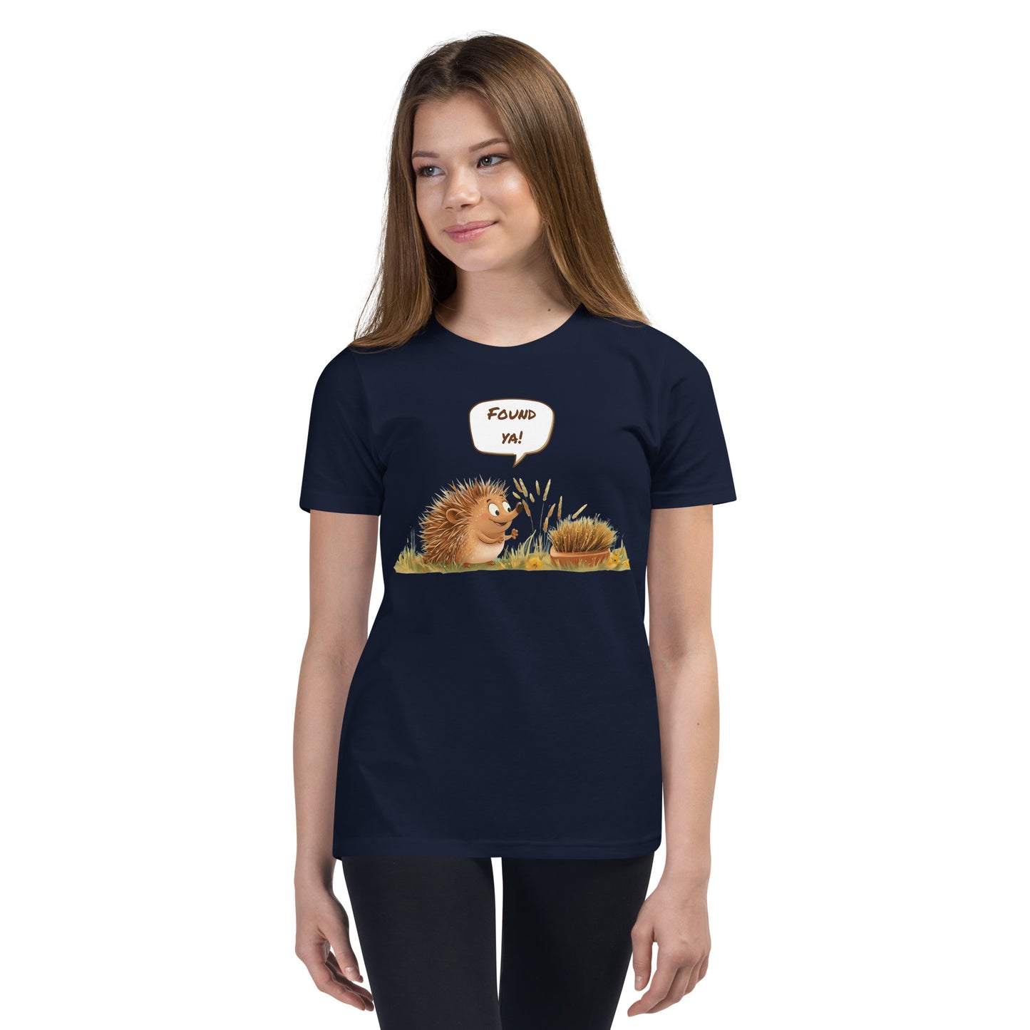 Product mockup. Ultra-soft premium youth navy blue t-shirt featuring cute cartoon hedgehog with speech bubble saying 'FOUND YA!' over grassy ground, in front of a brush, modeled on young girl.