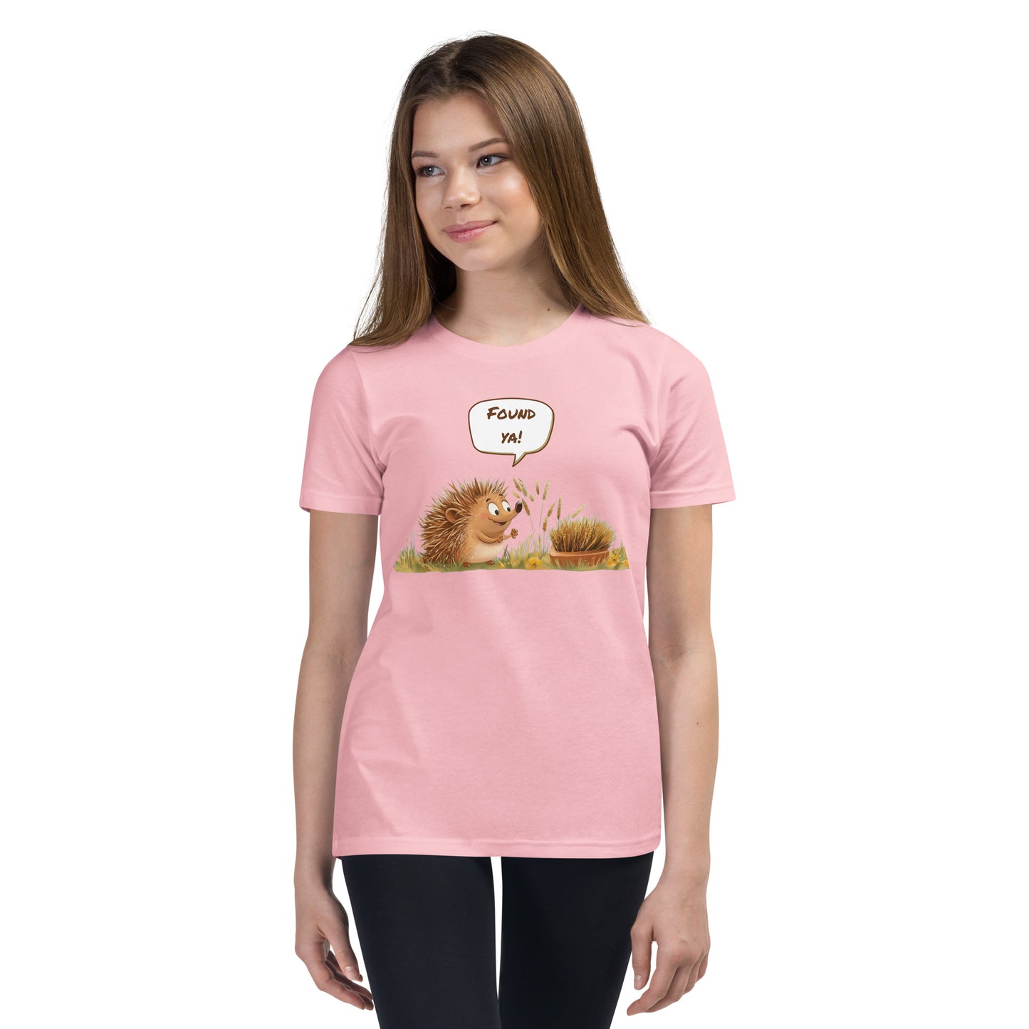 Product mockup. Ultra-soft premium youth pink t-shirt featuring cute cartoon hedgehog with speech bubble saying 'FOUND YA!' over grassy ground, in front of a brush, modeled on young girl.