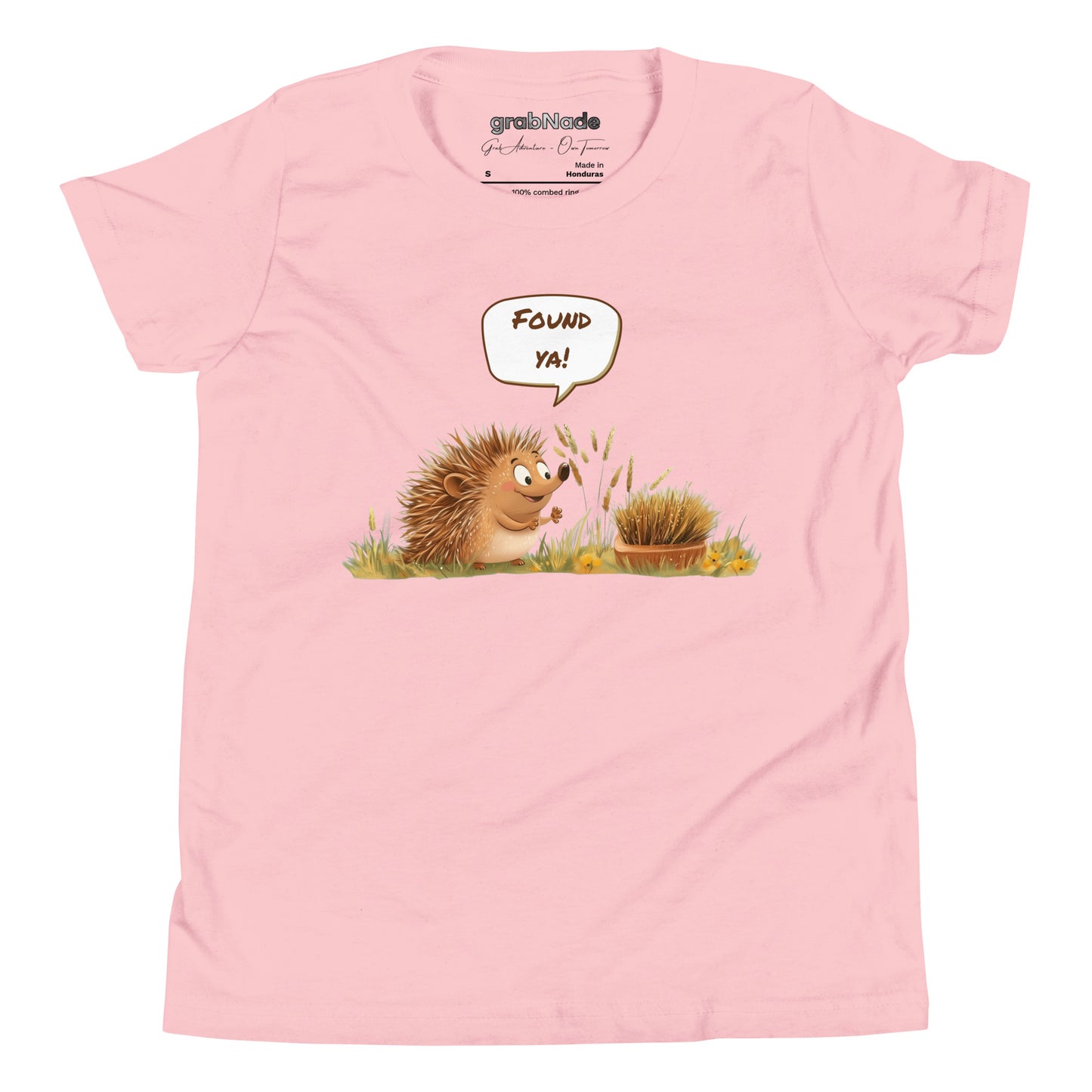 Product mockup. A smiling child wearing a light pink t-shirt printed with a playful illustration of a cheerful hedgehog popping up from the grass and saying “Found ya!” in a speech bubble, beside a second hedgehog curled into a ball, all set against a clean studio background.