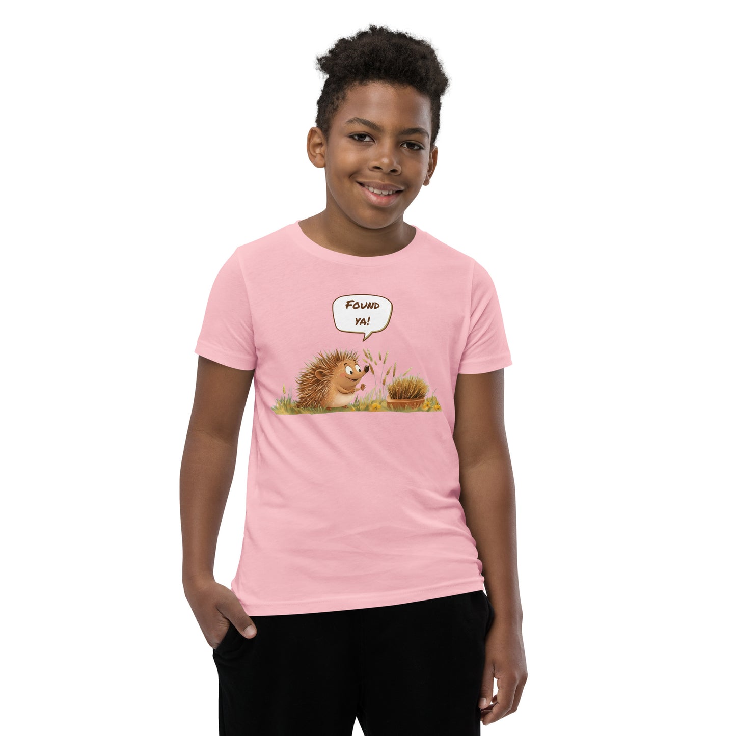 Product mockup. A smiling child wearing a light pink t-shirt printed with a playful illustration of a cheerful hedgehog popping up from the grass and saying “Found ya!” in a speech bubble, beside a second hedgehog curled into a ball, all set against a clean studio background.
