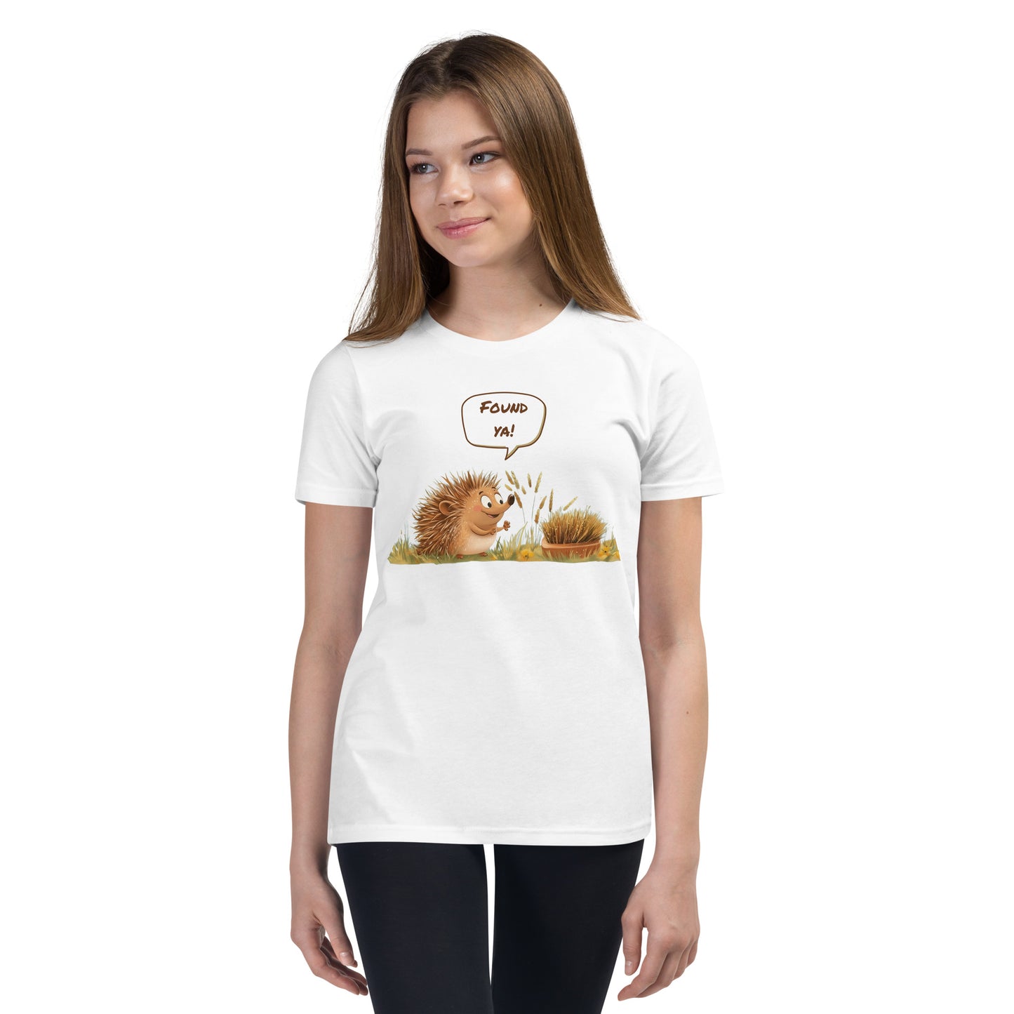 Product mockup. Ultra-soft premium youth white t-shirt featuring cute cartoon hedgehog with speech bubble saying 'FOUND YA!' over grassy ground, in front of a brush, modeled on young girl.